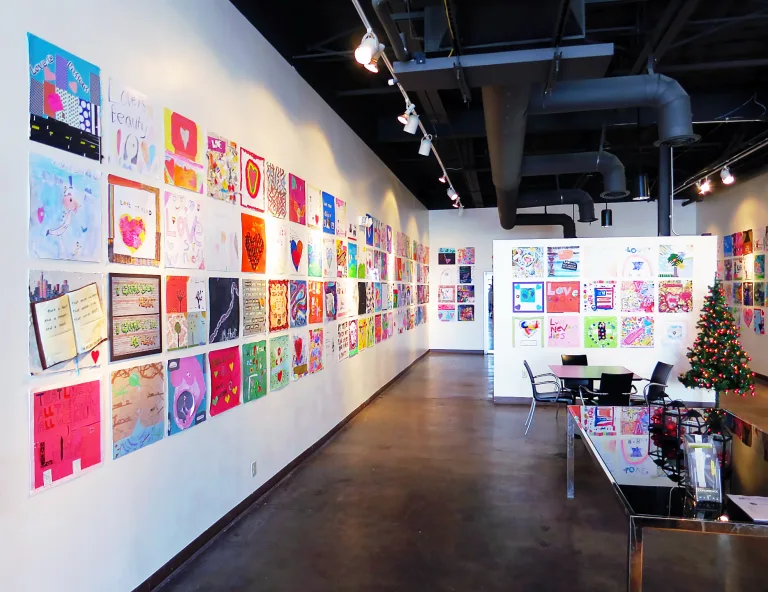 Art gallery with walls covered in colorful, love-themed children's artwork. A small decorated Christmas tree stands near a seating area.