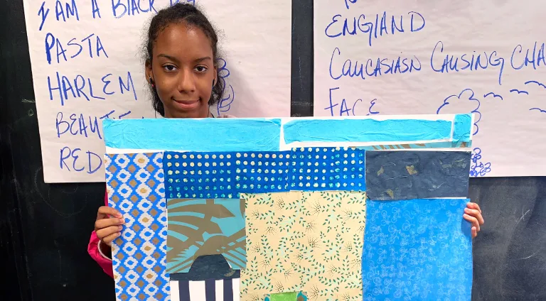 A young woman stands in front of a chalkboard with notes and holds up an abstract collage with multicolored patterned paper.