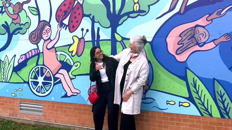 Two women hug and smile at each other while standing in front of the completed Burnet Elementary mural, which depicts children in a vibrant, whimsical nature scene.