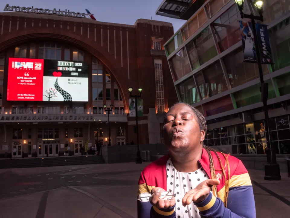 A woman poses while blowing a kiss outside the plaza entrance of the American Airlines Center. Behind her on the digital display is an illustration of a brick road leading to a heart, which is next to a tree and a quote by Haruki Murakami that reads “Love can rebuild the world.” in cut-out text. To the left is The American Love Project logo and the text “Art by Riley Berner, Lincoln, Nebraska."