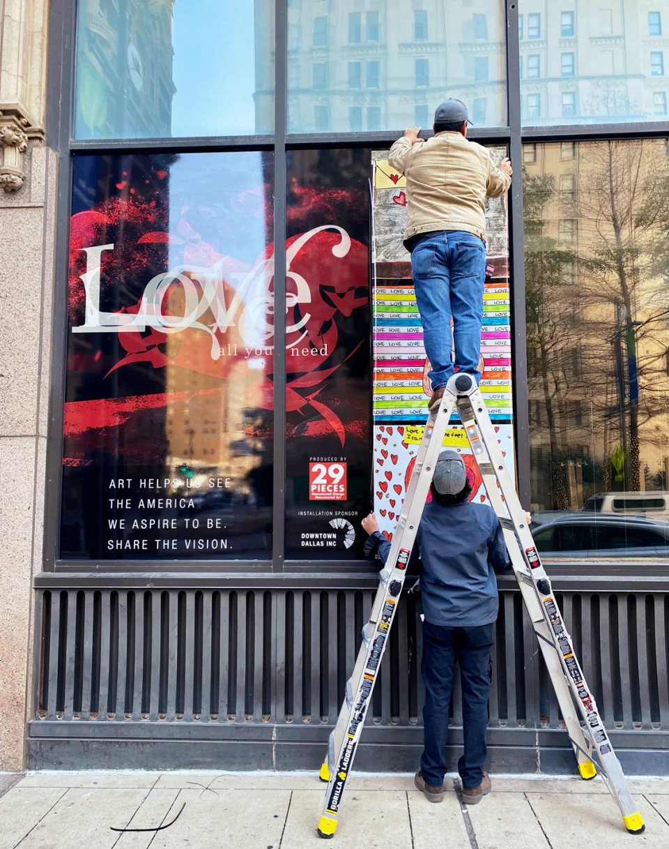 A man assists another man on a ladder who is installing a banner featuring American Love Project artwork in a storefront window display. In the bottom left of the installation is the text, “Art helps us see the America we aspire to be. Share the vision.” next to the 29 Pieces logo and the Downtown Dallas Inc. sponsor logo. 