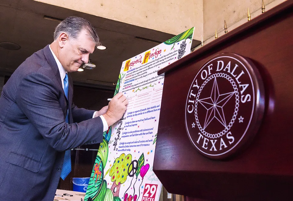 Rawlings signs a large poster board titled “Respect Pledge,” which is decorated with peace signs, hearts, trees and other shapes. In the foreground is a podium with the City of Dallas seal.