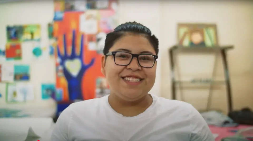A young woman wearing glasses smiles in a brightly lit room with art covering the wall.