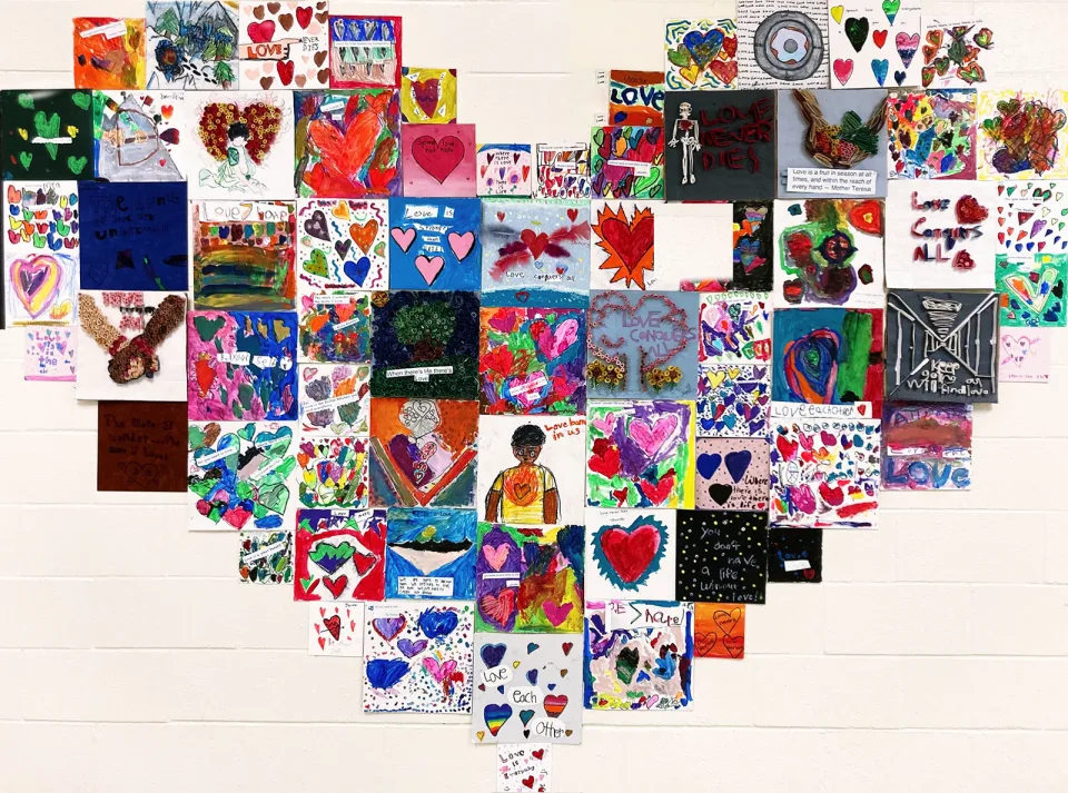 A concrete block wall is decorated with dozens of pieces of colorful student artwork arranged in the shape of a giant heart.