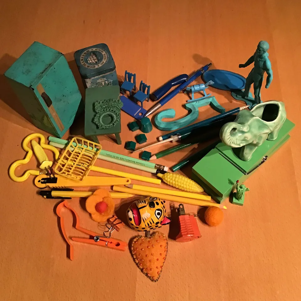 Bird's eye view of found objects, including pencils, clothes pins, a toy soldier, and a ceremic elephant, are arranged in the shape of a heart and sorted in rainbow color order.