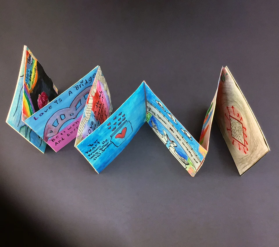 Bird's-eye view shot of a handmade accordion-style book, made up of square pages that each contain a different drawing and quote.