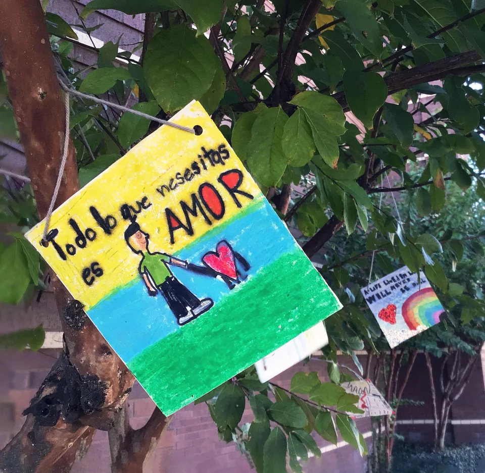 Colorful student artwork, including a drawing of a boy holding the hand of a stick figure heart with the text “All I need is love” written in Spanish, hangs from trees outside a school.