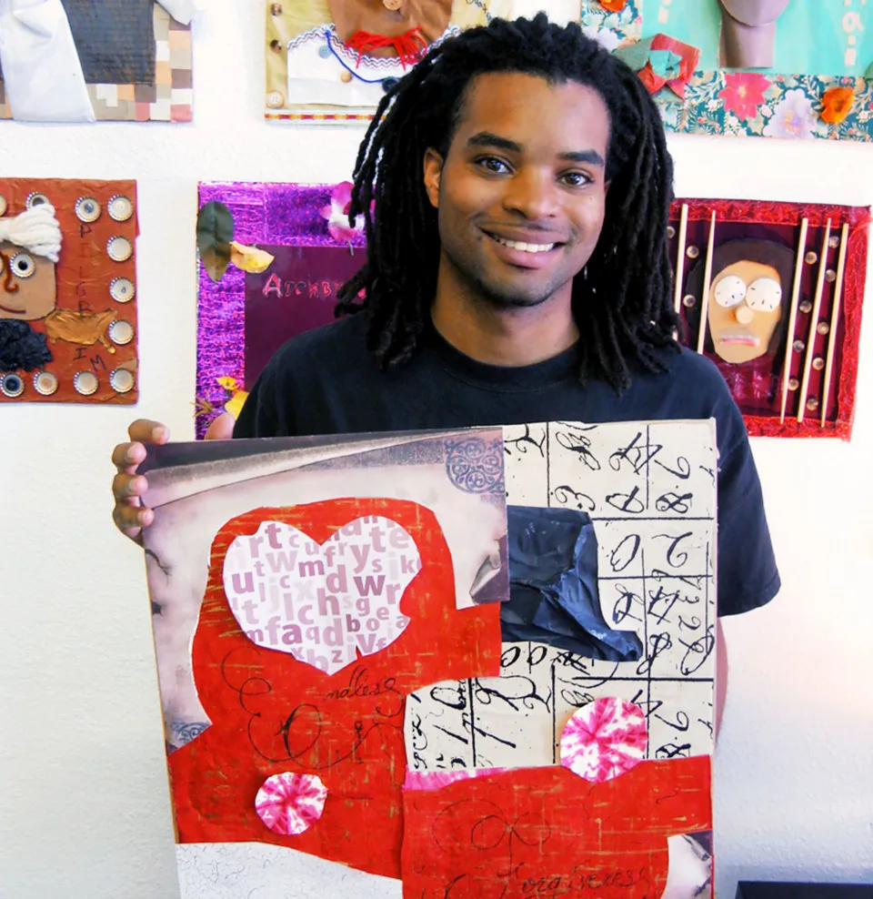 A young man smiles holds up a collage with a heart and abstract shapes and patterns while standing in front of a wall covered in art.