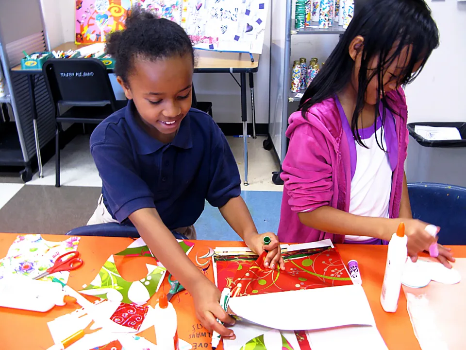 A young girl and a boy work on an art project together at a table with scraps of paper, glue and scissors.