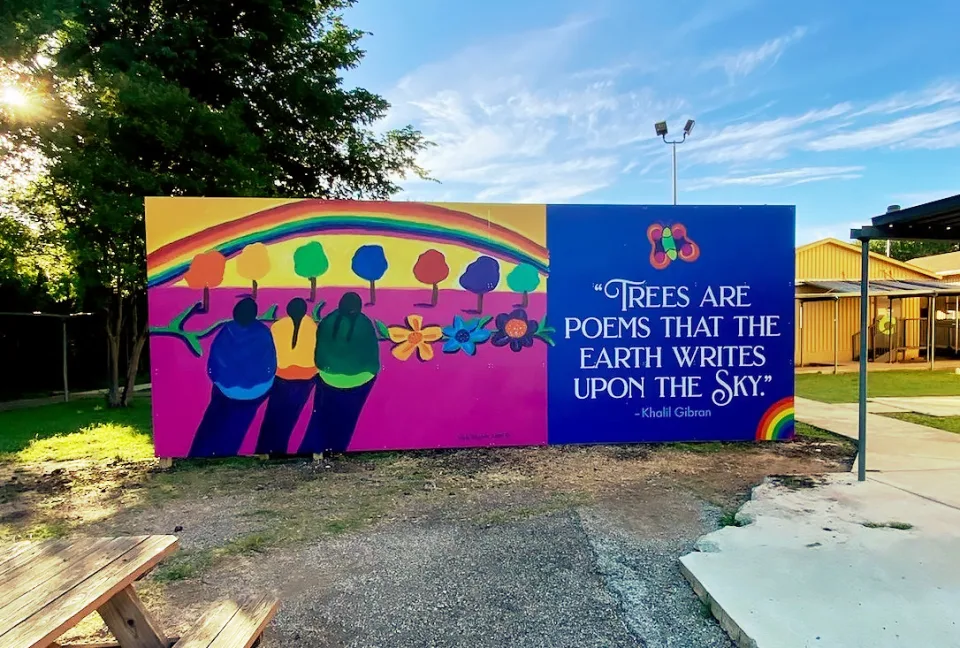 Colorful mural showing three figures facing a rainbow and a row trees. Beside them is a quote by the writer Khalil Gibran that reads, “Trees are poems that the earth writes upon the sky”.
