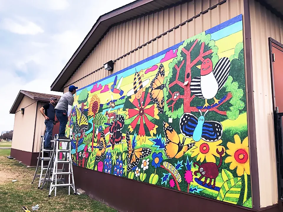Thomas Printworks crew members stand on a ladder while installing a vibrant mural, which depicts a busy nature scene with flowers, butterflies and birds, on the side of a building on the John Ireland Elementary School campus.