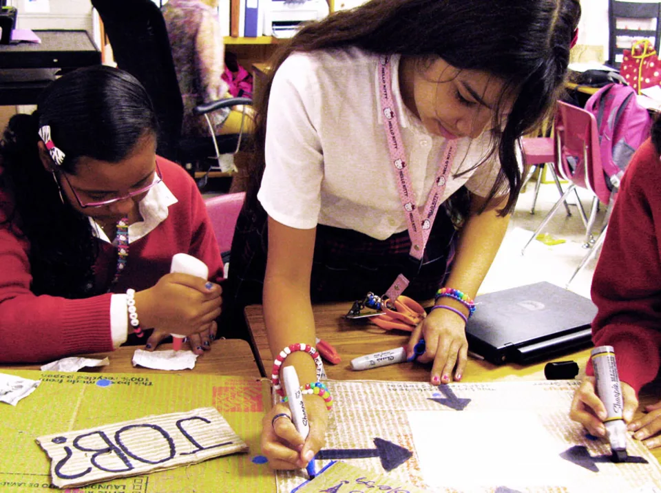 Students work intently on an art project in a classroom, using Sharpies and Elmer’s glue to draw arrows and attach pieces of paper to a poster board with the text “Job?”.