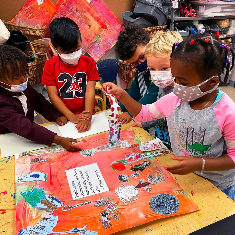 A group of elementary-aged students stands around a table and attaches cutout drawings to a collage, which has an Ojibwa prayer in the center.